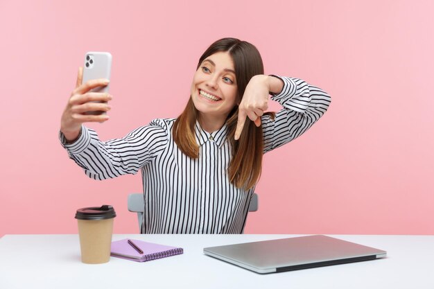 Cute positive woman blogger in striped shirt pointing finger down recording video for followers, asking to subscribe and turn on notifications. Indoor studio shot isolated on pink background