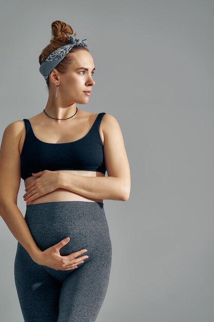 Cute pregnant belly isolated on gray Side view of young pregnant woman embracing her abdomen with hands Big belly on the third trimester of pregnancy closeup Concept of pregnant life