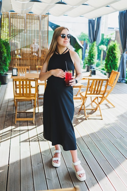 Photo a cute pregnant woman in a black dress on a summer terrace in a cafe drinks fresh lemonade a pregnant woman drinks nonalcoholic fruit lemonade