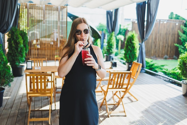 Photo a cute pregnant woman in a black dress on a summer terrace in a cafe drinks fresh lemonade a pregnant woman drinks nonalcoholic fruit lemonade