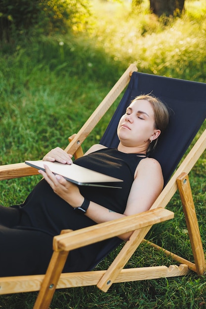 A cute pregnant woman is sitting in a park and working remotely on a laptop Online work on maternity leave Modern pregnant woman