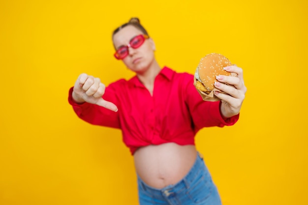 A cute pregnant woman in a pink shirt and pink glasses eats fast food a pregnant woman on a yellow background with an appetizing burger in her hands harmful food for pregnant women