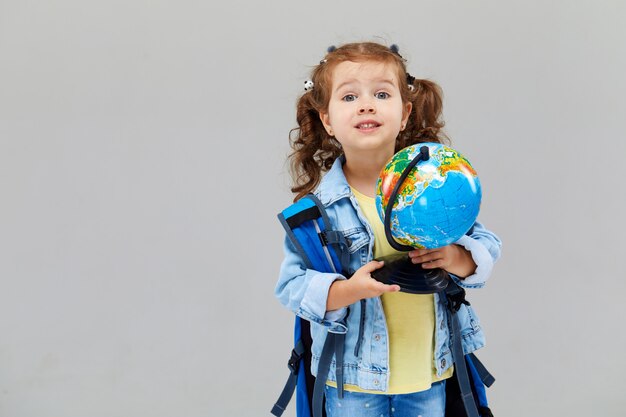 Cute preschool girl with a blue backpack on her back, holding a globe in her hands. 