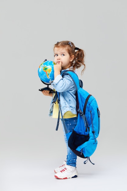 Cute preschool girl with a blue backpack on her back, holding a globe in her hands.