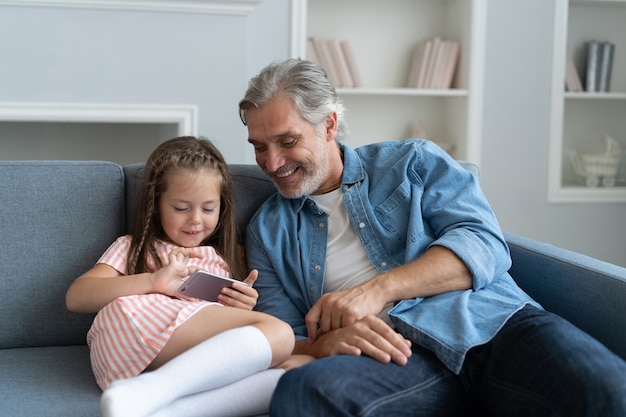 Милая дошкольница помогает отцу показать что-то на смартфоне, умная маленькая дочь и папа сидят на диване и держат мобильный телефон