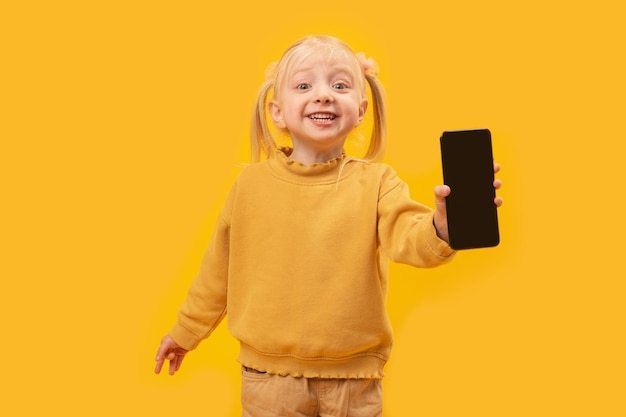 Cute preschooler little blonde girl stand on yellow studio background and hold smartphone Kid shows black phone with blank screen