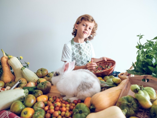 Cute preteen boy cooking vegan salad in bowl on kitchen counter with fresh vegetables and white fluffy rabbit and looking at camera