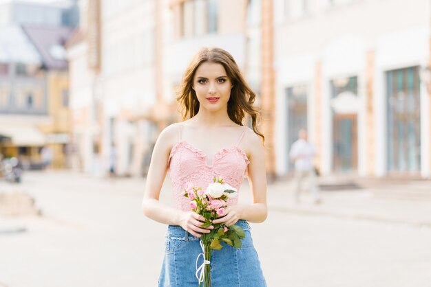 Cute pretty girl with professional makeup in a pink top in a summer city holding a bouquet of white and pink roses