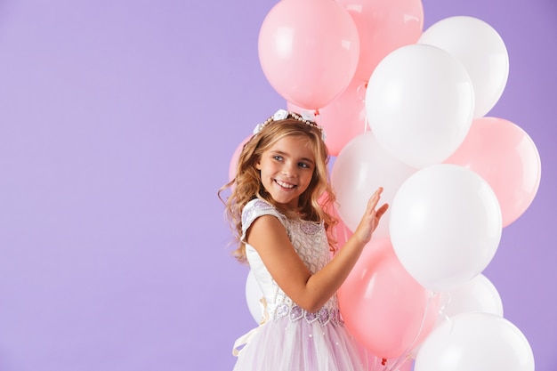 Cute pretty little girl dressed in princess dress isolated over violet wall, holding bunch of balloons