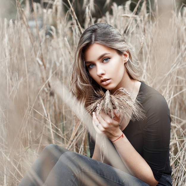 Милая симпатичная сельская молодая женщина с круглыми глазами в винтажной черной футболке и модных джинсах отдыхает, сидя на открытом воздухе в сухой осенней траве
