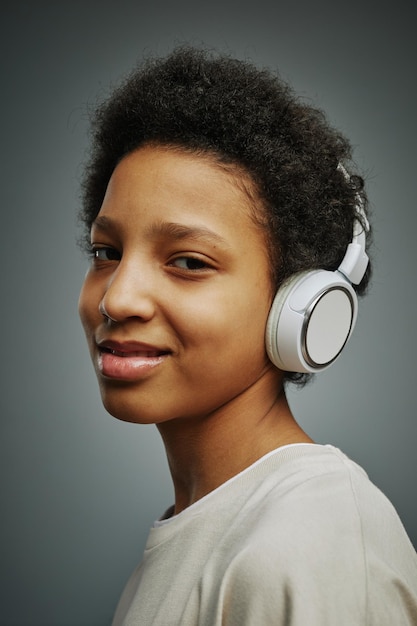 Cute pretty youthful girl listening to music in headphones and looking at camera while standing over