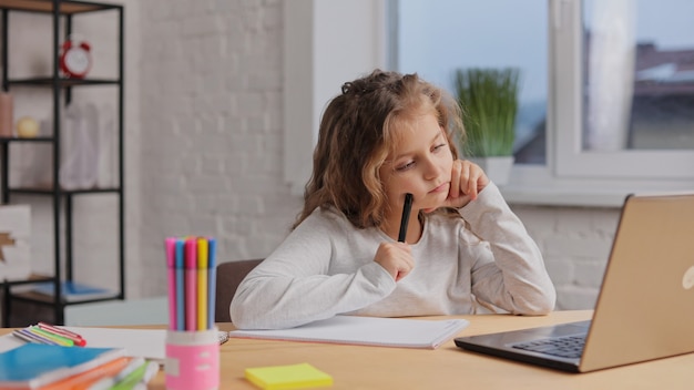 Милая девочка начальной школы учится дома, используя портативный компьютер. У школьницы онлайн-урок. Дистанционное обучение, домашнее обучение