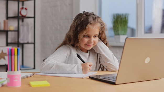 Cute primary school girl studying at home using laptop computer. Schoolgirl has online lesson.