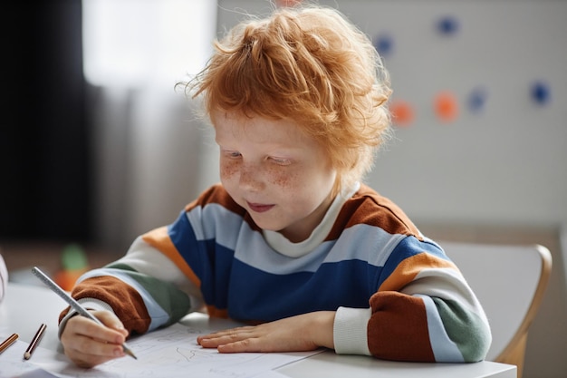 Cute primary school learner with freckles and ginger hair drawing with pencil while sitting by table