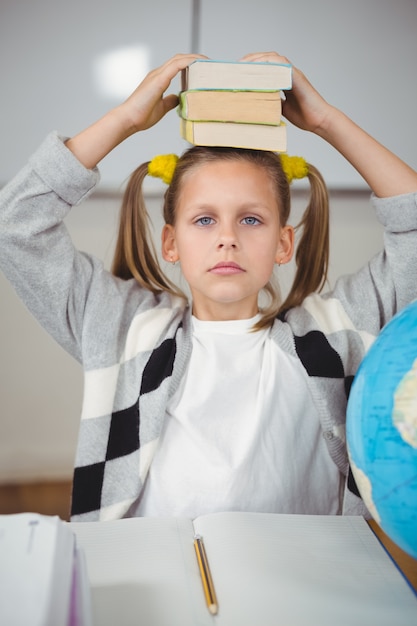 Cute pupil balancing books on head in a classroom