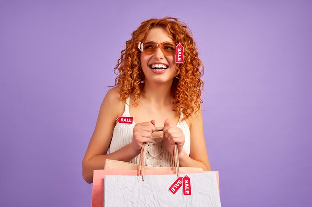 Cute red-haired girl with curls and shopping bags and sale labels isolated on purple