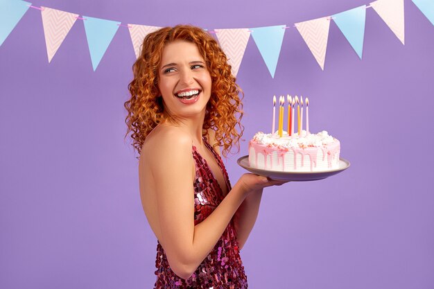 Cute red-haired woman with curls in a shiny dress holding a cake with candles isolated on purple