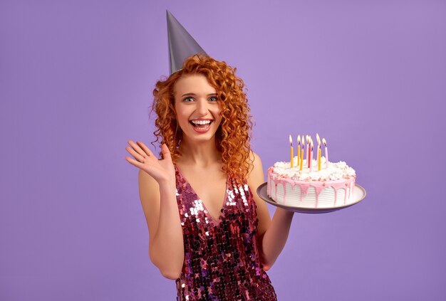 Cute red-haired woman with curls in a shiny dress and a party cap is holding a cake isolated on purple