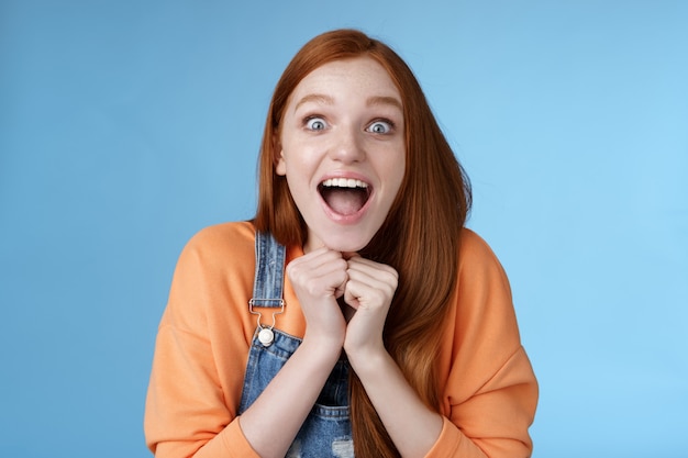 Cute redhead european girl blue eyes freckles reacting amused shocking rumor lift eyebrows drop jaw surprised smiling excited picked get role theatre play rejoicing astonished blue background