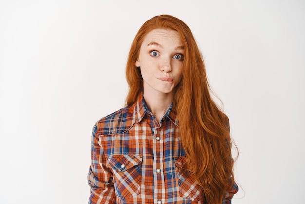 Photo cute redhead girl standing indecisive over white background sucking lips and staring at camera surprised