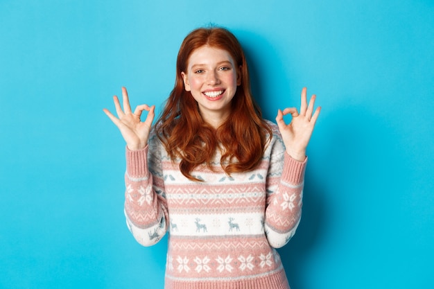 Cute redhead girl in sweater showing okay signs, smiling pleased, approve and like, praise excellent choice, standing over blue background.