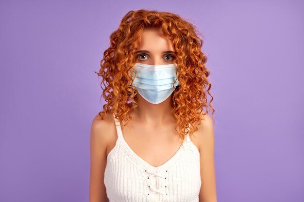 Cute redhead girl with curls in protective medical mask isolated on purple wall