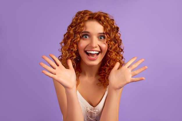 Cute redhead girl with curls shows emotion of surprise, shocked isolated on purple wall