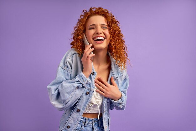 Cute redhead hipster girl with curls in a denim jacket talking on the phone isolated on a purple wall