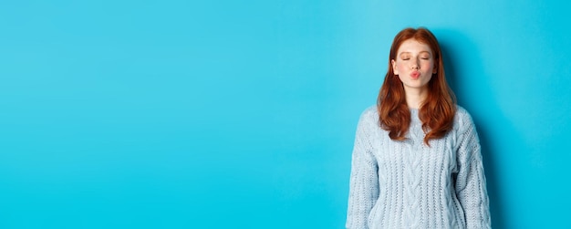 Cute redhead teen girl waiting for kiss pucker lips and close eyes standing in sweater against blue