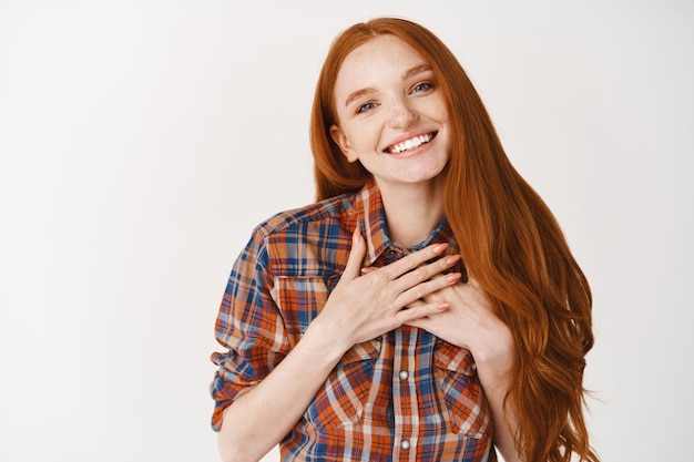 Cute redhead woman with pale skin and blue eyes, holding hands on heart and smiling thankful