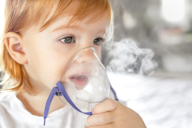 Cute redheaded twoyearold child breathing through nebulizer mask sitting at home side view with text space closeup