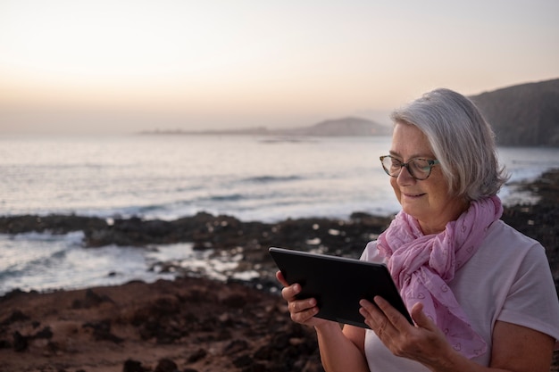 Cute relaxed senior woman gray hair sitting near the beach surfing the net with tablet. Dusk light. Mountains and sea in background.