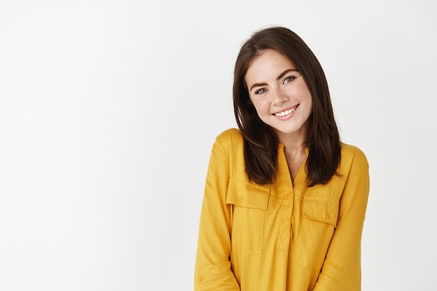 cute and romantic woman glancing at camera, smiling and looking shy, standing over white wall
