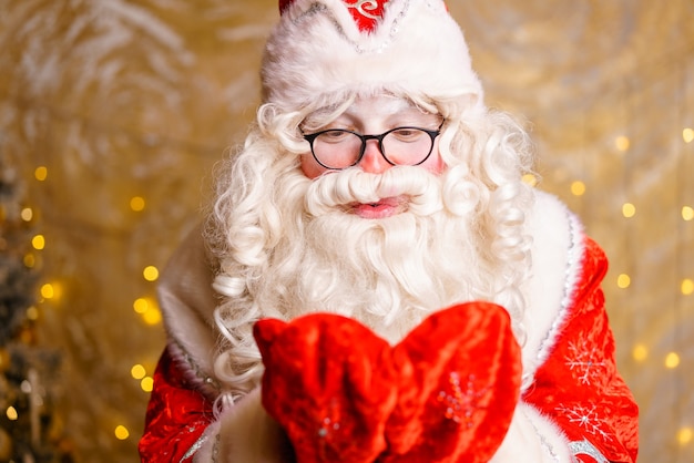 Photo cute santa claus in glasses on a wall background with a bright garland bokeh hands in mittens closeu...