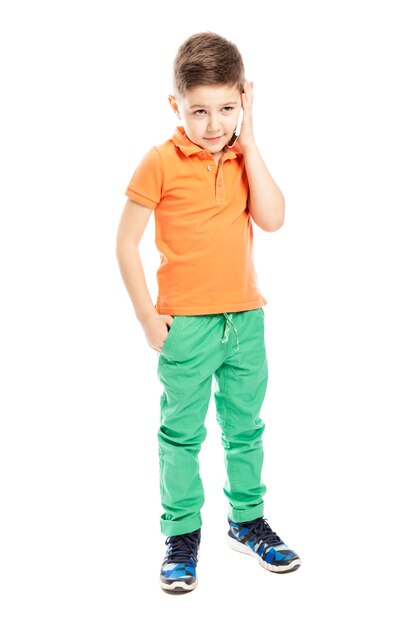 A cute school-age boy in a bright orange polo t-shirt is talking on a smartphone. Full height. Vertical. Isolated on a white background.