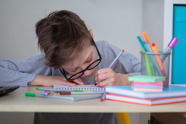 机に座ってメモを書いて家で勉強している眼鏡のかわいい学校の生徒の子供の男の子。