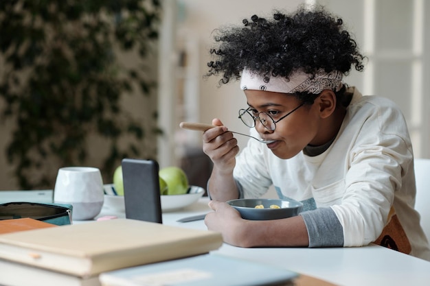 Photo cute schoolboy eating muesli for breakfast and watching online video