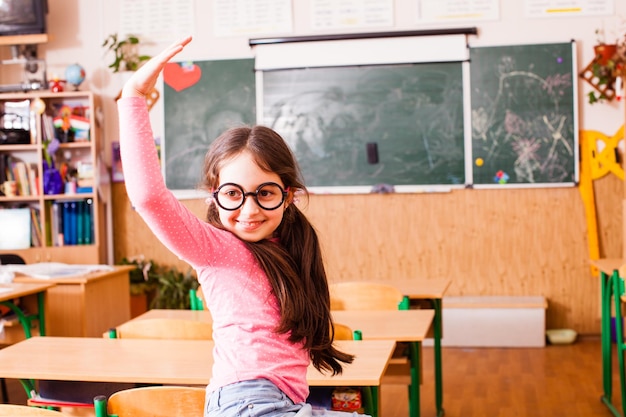 Cute schoolgirl in funny big glasses, sitting on the desktop and smiling