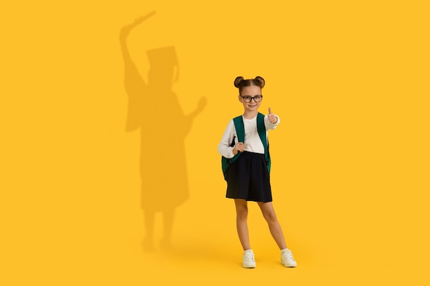 Cute Schoolgirl Showing Thumb Up Having Shadow In Graduation Costume On Background