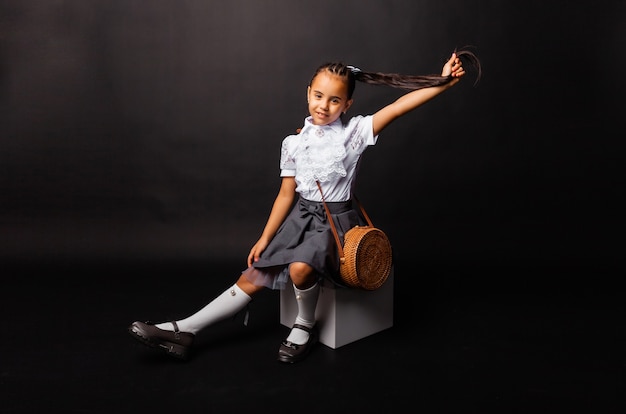 Cute schoolgirl sitting on a white cube and holding a wicker round bag, studio portrait on a dark background