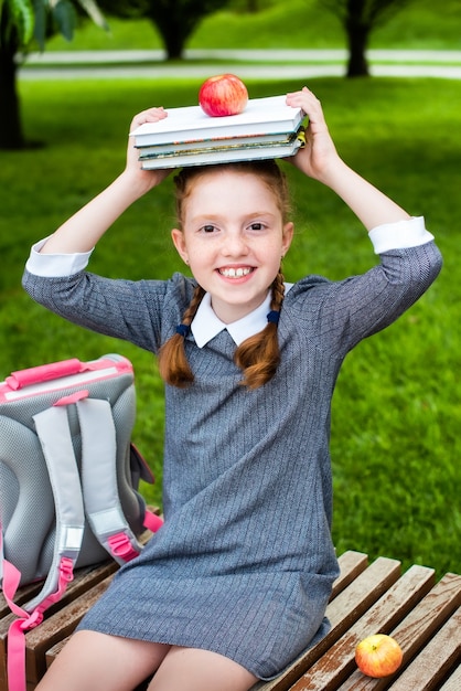 Cute schoolgirl smiling with a book stack and an apple over her head sitting in the park