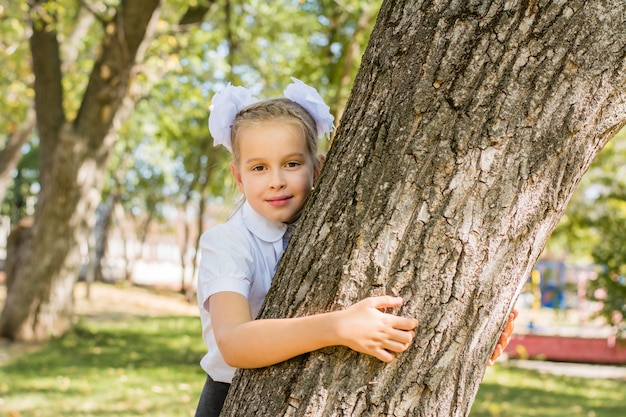 하얀 리본을 단 귀여운 여학생이 화창한 가을 공원의 나무 옆에 서 있습니다. 9월 1일 1학년. 학교 개념으로 돌아가기
