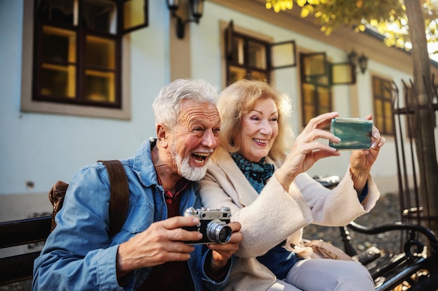 Cute senior couple sitting on the bench and taking a selfie. Man is holding camera while woman holding a cell phone.