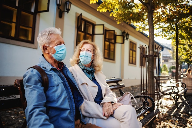 外のベンチに座っておしゃべりに保護マスクを付けたかわいい年配のカップル。