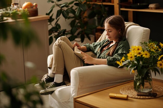 Cute serious brunette girl with pigtails sitting in armchair and reading book