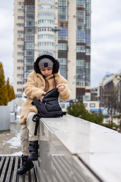 彼女の手にバックパックを持ったかわいい7歳の女の子が街の欄干に座っています