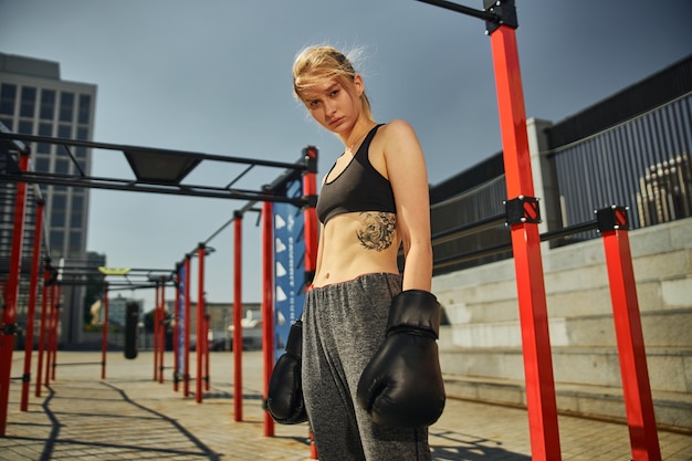 Cute shot of young woman in black boxing gloves who is starting her kickboxing training on the sport ground