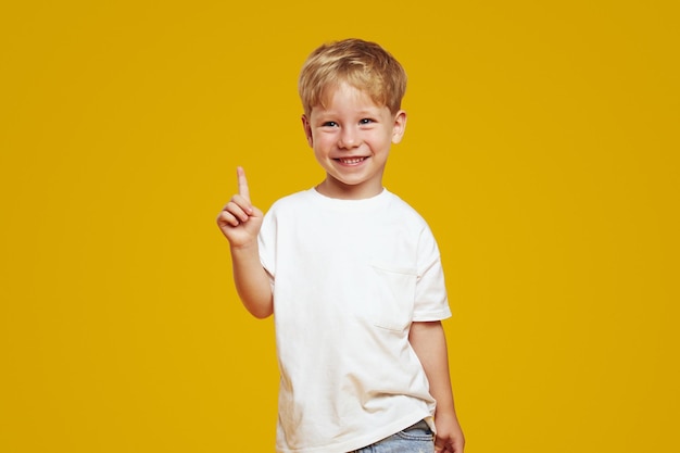 Foto piccolo ragazzo biondo e timido che indossa una maglietta bianca sorridendo e indicando verso l'alto mentre guarda lontano