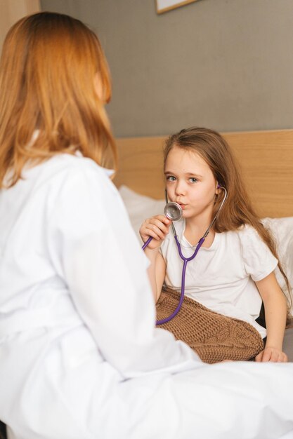 Cute sick little girl playing with stethoscope while lying in bed in bedroom during visit of pediatrician at medical clinic