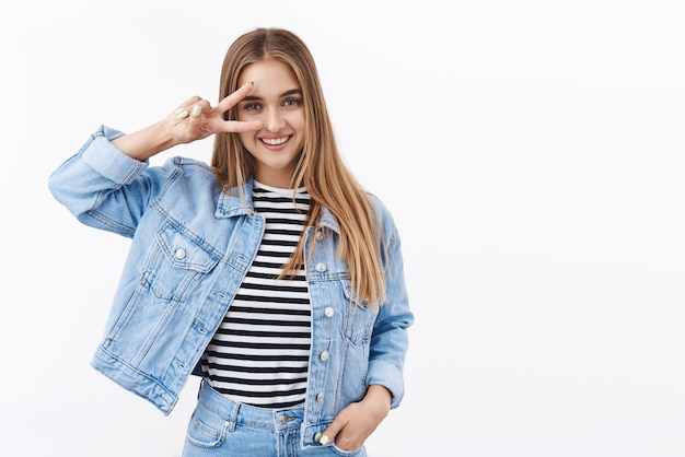 Cute and silly blond caucasian 25s girl in denim clothes show kawaii peace gesture over eye and smiling ready standing joyful express positivity and happy emotions white background